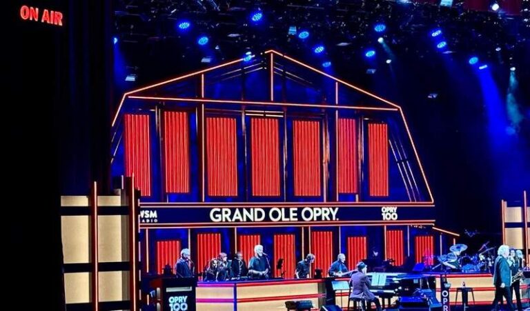 An outline of the Grand Ole Opry in lights fills part of the music hall stage. Blocks of light change colors from red to blue to green. Singers and musicians perform in front of the lighted image.