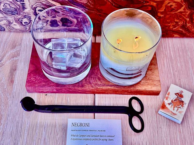 Two matching short clear glasses sit side by side. One holds a candle, the other some chunky ice cubes. The candle scent matches the cocktail recipe provided with the ice cube glass.