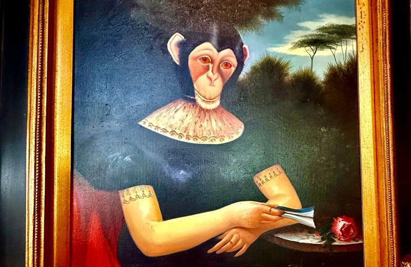 A monkey wearing a dark green dress with short sleeves and a wide lace collar sits at a small table. She is a framed portrait seeming to be outside under a blue sky and next to tall trees. 