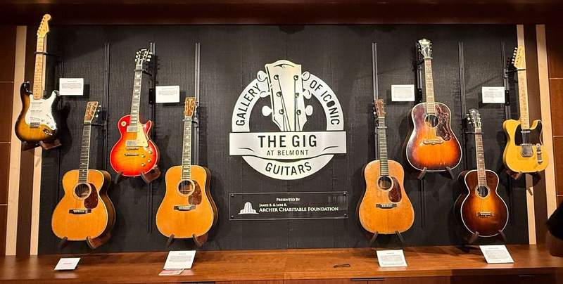 Eight guitars hang longways on a black wall with the words Gallery of Iconic Guitars in the middle. Small labels next to each guitar tells its history.