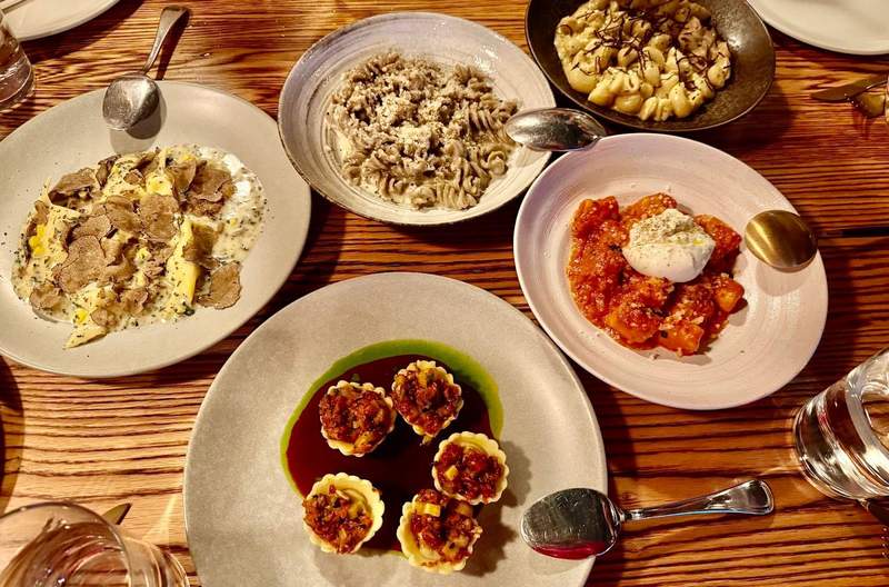 Five plates and wide shallow bowls hold handmade pasta dishes to share with tablemates. A serving spoon on each dish rests on the edge.The table is wood with no cloth. 