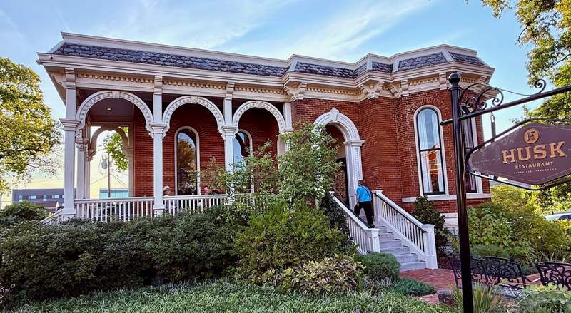 Husk Restaurant is a horizontal building, red brick with white trim along the roof and porch and around the long vertical windows. The yard is lush with green shrubs and lawn.