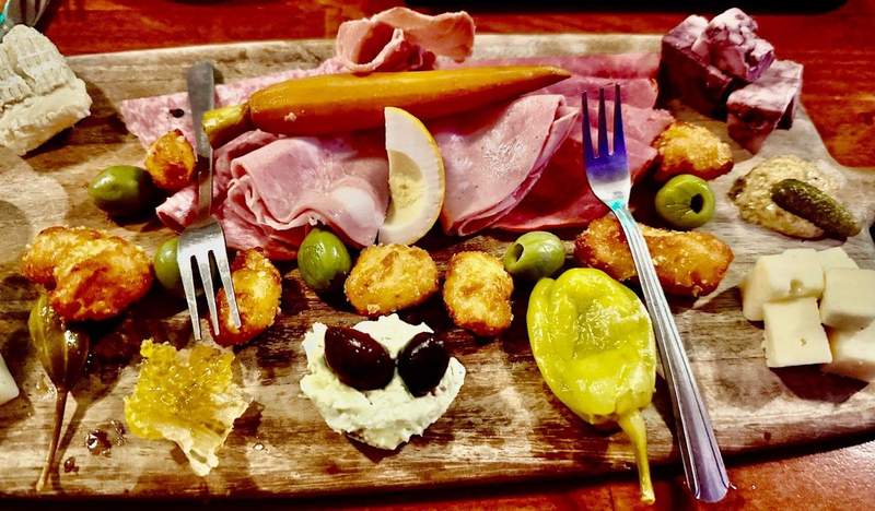The wooden charcuterie board is visible only around its edges -- filled with bite-sized pink meats and white and yellow cheeses. Olives are black or green, peppers and pickles shades of green. 