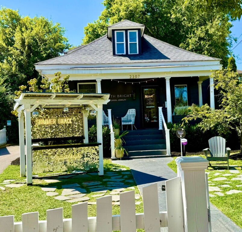 Shimmer Shack are the glittery words on the front yard sign of a former residence now housing a jewelry store. Green trees, blue sky and a white picket fence surround the store which is dark blue, trimmed in white.