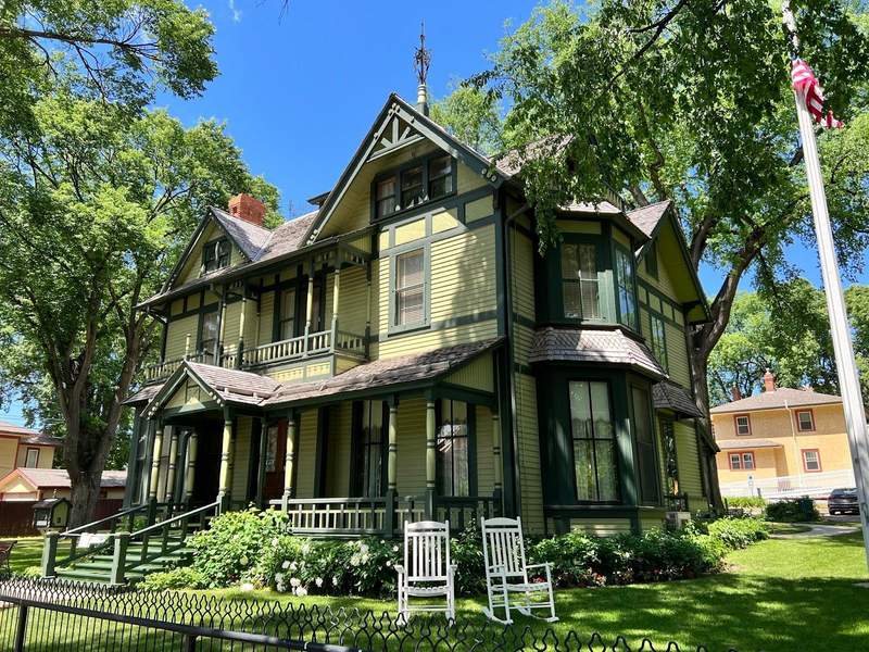 A pretty Victorian house painted in shades of green a wide front porch and upper balconies. Two white wooden rocking chairs in front of home.
