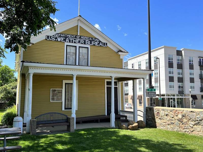 A small vintage yellow wooden building with a porch supported by slim columns. the sign reads Department of Agriculture U.S. Weather Bureau.