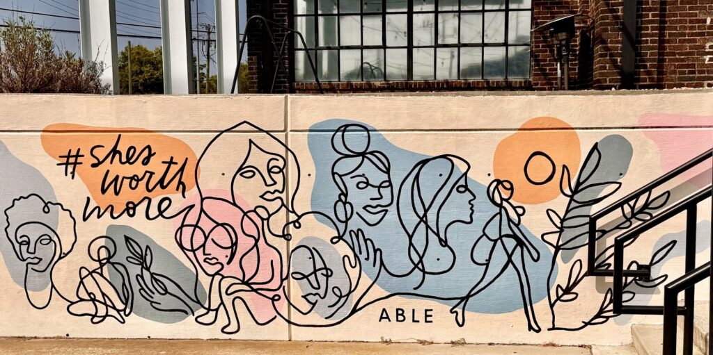The long narrow mural on the sidewalk outside ABLE store shows faces and some full body views of women, drawn with simple lines. The words she's worth more in the upper left corner have a hashtag symbol. Colors are just splashes of pale blue, pink and salmon.