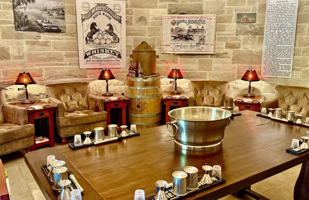 The walls are horizontal stone blocks. Chairs along the walls are padded and soft, each with a small table and short lamp. In the center is a long wooden table with sets of four pewter tasting cups. Posters of horse racing and whiskey line the stone walls.
