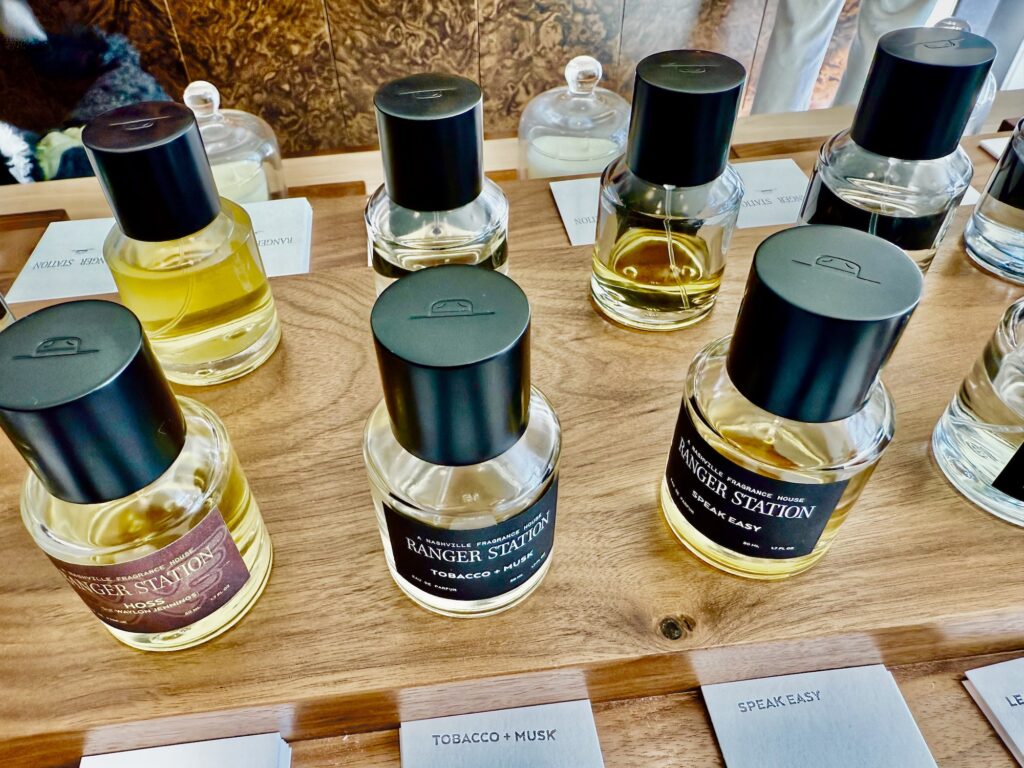 Seven look-alike small glass bottles with Ranger Station labels and dark green lids line up on a wooden table. Each has a separate label describing the ingredients of the hand-crafted scent. Many more are further down the table.