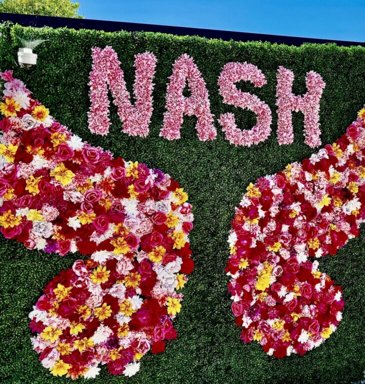 The vertical garden wall has artificial dark green grass as the background and the capital letters NASH in solid pink across the top. Big wings in pink, red, yellow and white flowers leave space for a person to stand and be photographed.