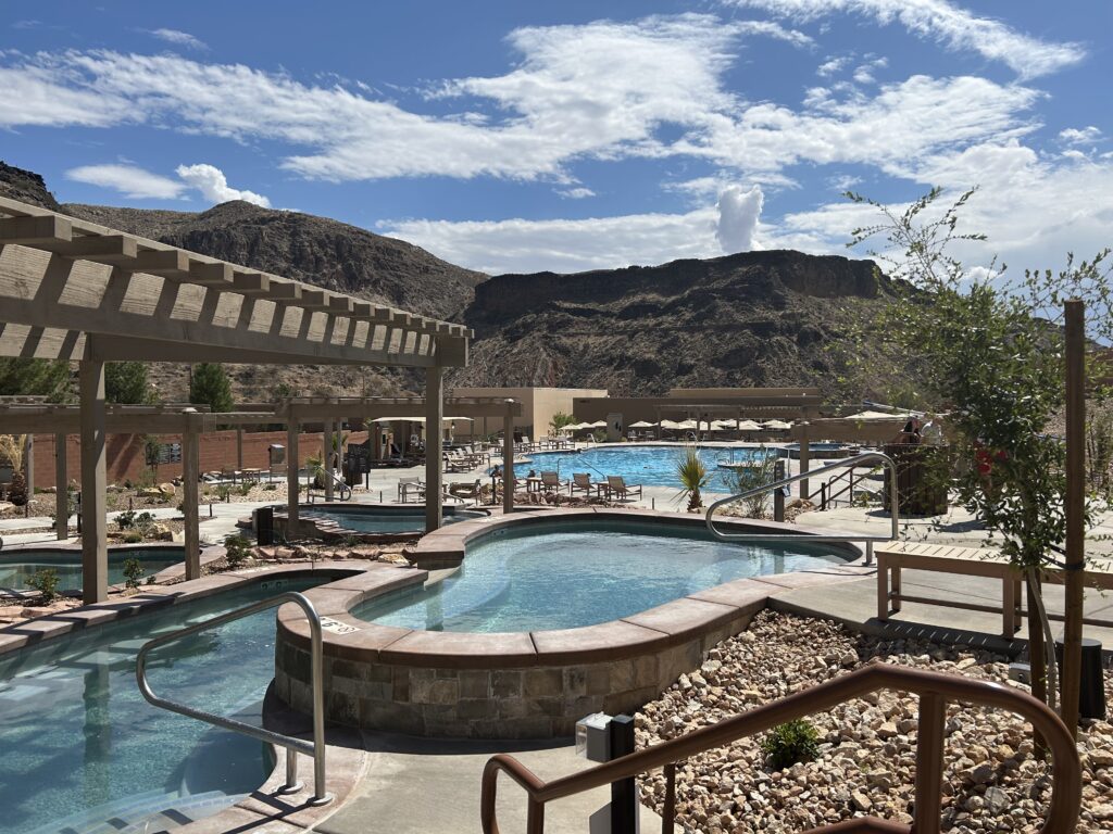 Zion Canyon Hot Springs is filled with numerous pools to dip into each with different temperatures and mineral make-up. See a few of the pools with turquoise water and rocks in the distance under blue skies with fluffy white clouds.