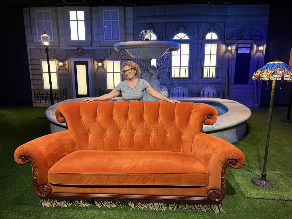 The author, wearing glasses and a grey top, is standing behind the famous orange couch from the opening scene of every "Friends" episode at a new must-see Las Vegas attraction. Behind her is the famous fountain and a large building with many windows. To her right is a Tiffany-like standing lamp.