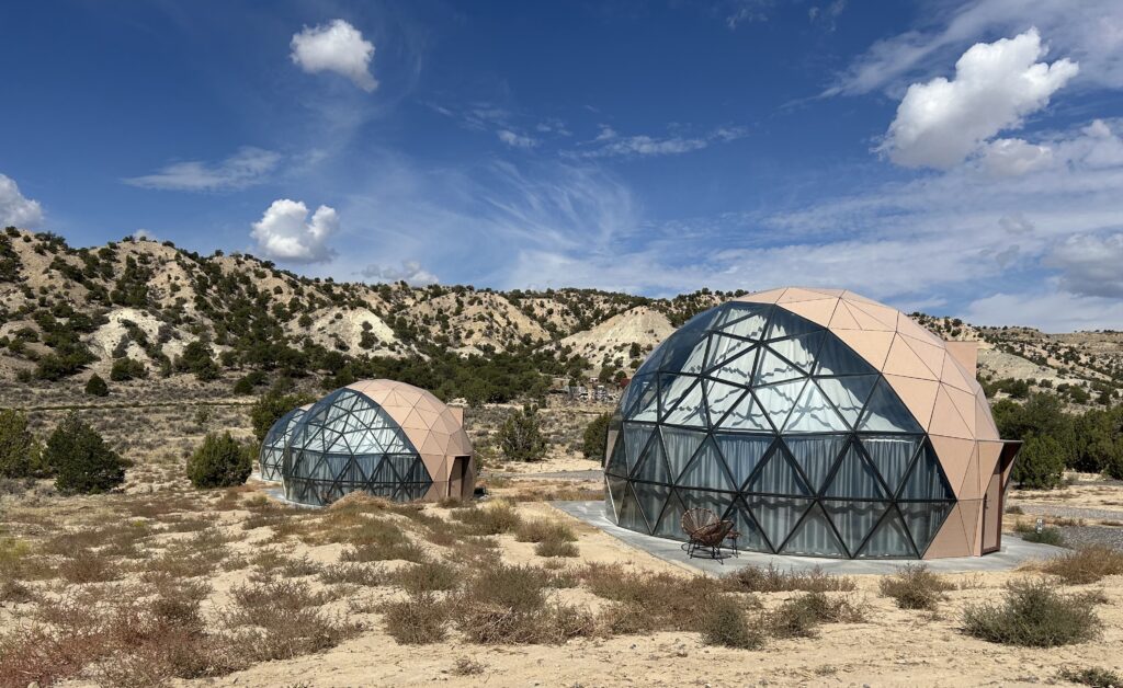 The glass domes at Clear Sky Resorts Bryce Canyon give you a great view of the night sky. Three of the geodesic domes that are hotel rooms. Curtains are closed but at night you can open them to see the sky and stars. There is a brown desert like landscape around them and blue sky above.