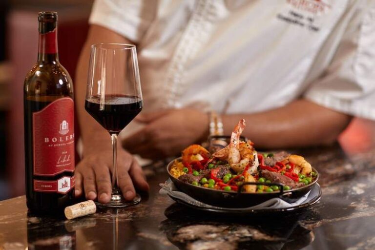 A glass of red wine and wine bottle sit next to a pan of paella with a chef in the background at Bolero Restaurante in Temecula.