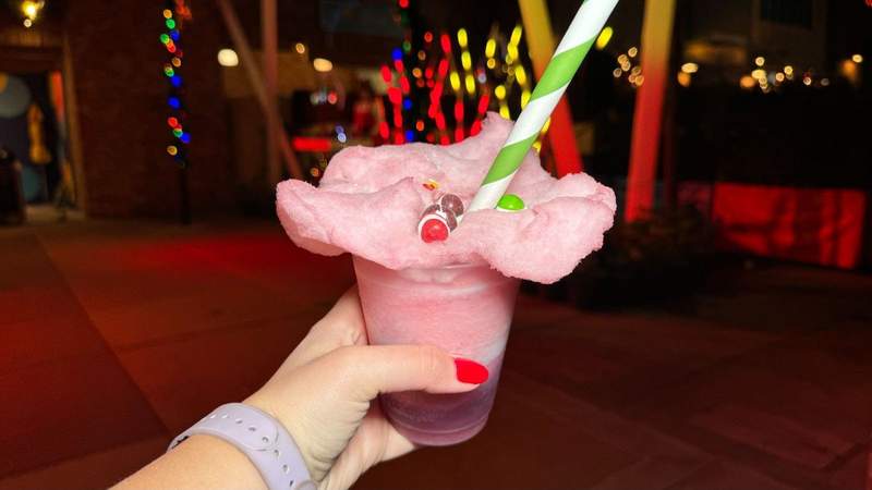 Hand holidng a cup with pink and purple non alcoholic beverage topped pink with cotton candy against a backdrop of lights.