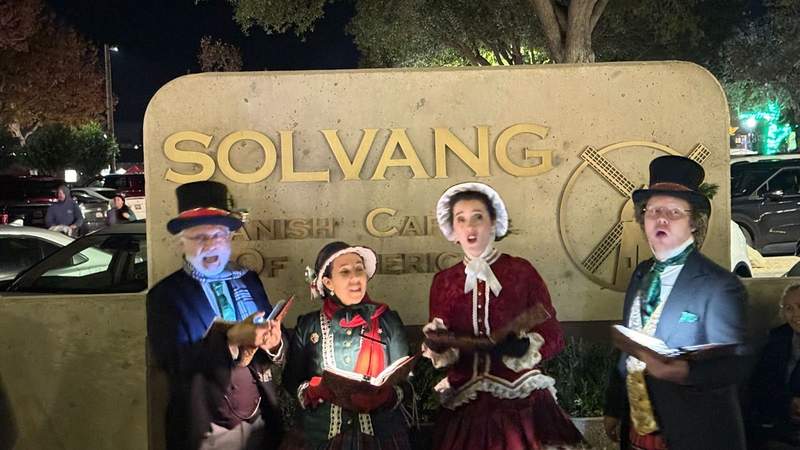 four people singing in Dickens themed outfits with open books infront of the Solvang sign during Solvang Julefest