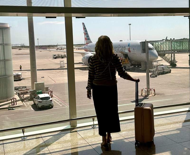woman with a carry-on suitcase looking out an airport window before a trip