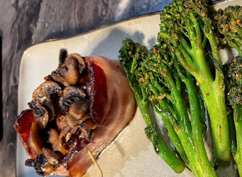 At Curries in Pine Bluff, Wyoming, a piece of bacon wrapped around a steak filet that is covered in mushrooms. Next to it is broccoli rabe.