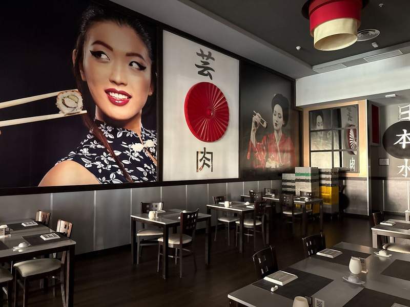 The image shows the colorful interior decor of the Tengoku Japanese Restaurant at Hotel Riu Palace. It shows tables set for patrons and pictures on the wall of attractive Japanese women and sushi.