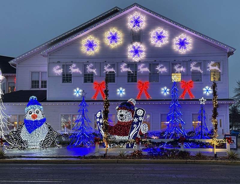 Lighted snowflakes reindeer bows trees and animals in front of a building.