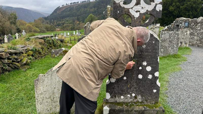 man wiping down a grave stone with grass at a cemetery.  