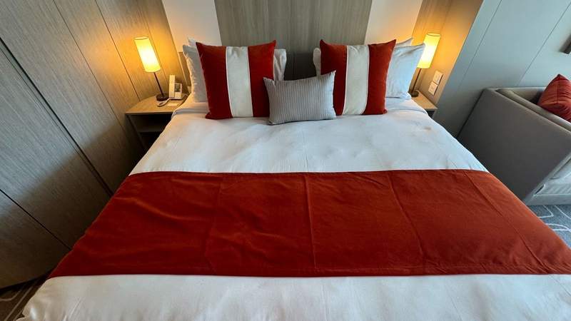 white bed with red bed runner and throw pillows with two night lights and a wooden closet in a stateroom on the celebrity xcel