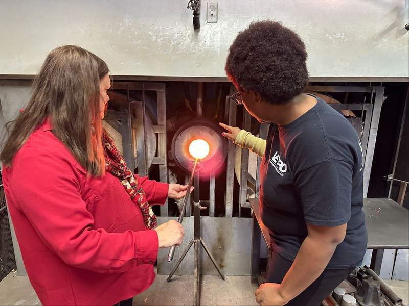 A woman heating a blob of glass and another person providing instructions.