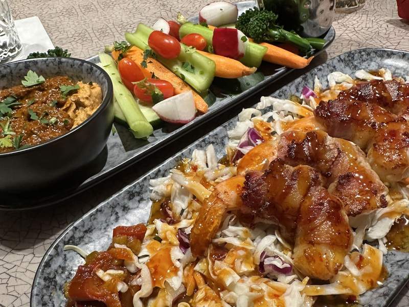 Two plates. One has a variety of raw veggies and a bowl of hummus. The other plate has bacon wrapped shrimp on cole slaw.