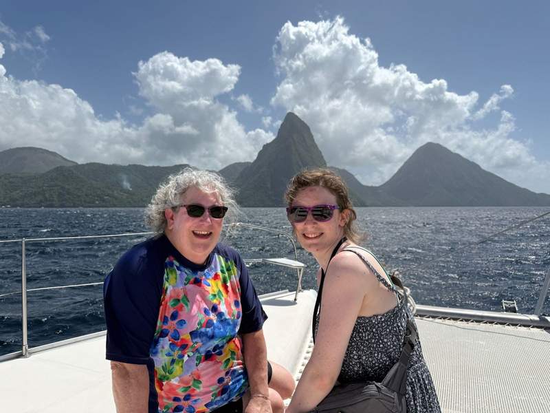 Two women in sunglasses look toward the camera on a boat deck with the Pitons of St Lucia in the background.