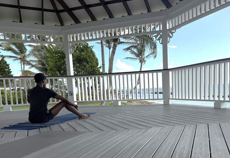 A man sits on a yoga mat facing the ocean in a white gazebo.