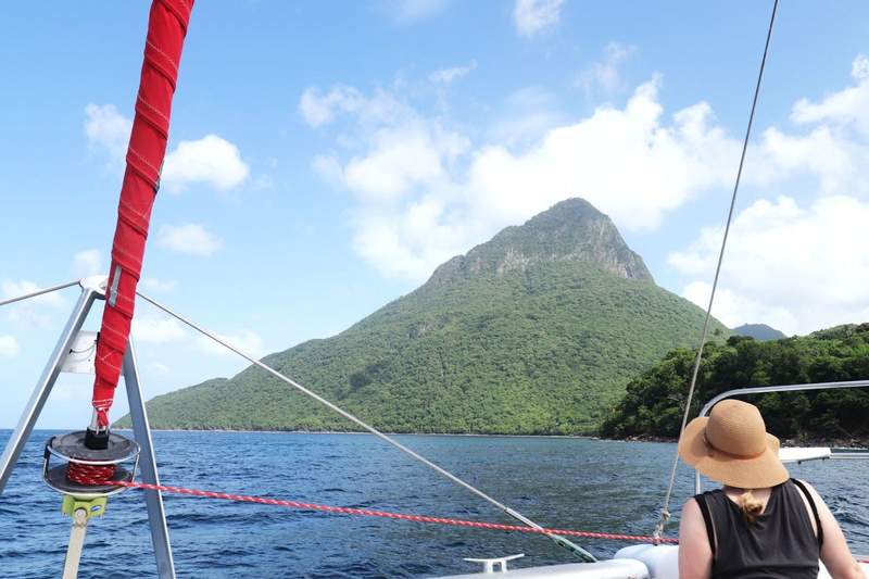 View from front deck of a catamaran of the PItons in St Lucia. A woman in black with a straw hat looks ahead through the boats rigging.