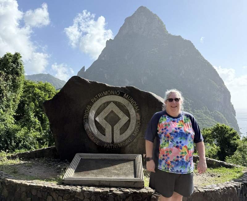 Woman in a bright floral top and black shorts stands in front of the sign designating the UNESCO World Heritage Site in St Lucia. Behind her is one of the Pitons.