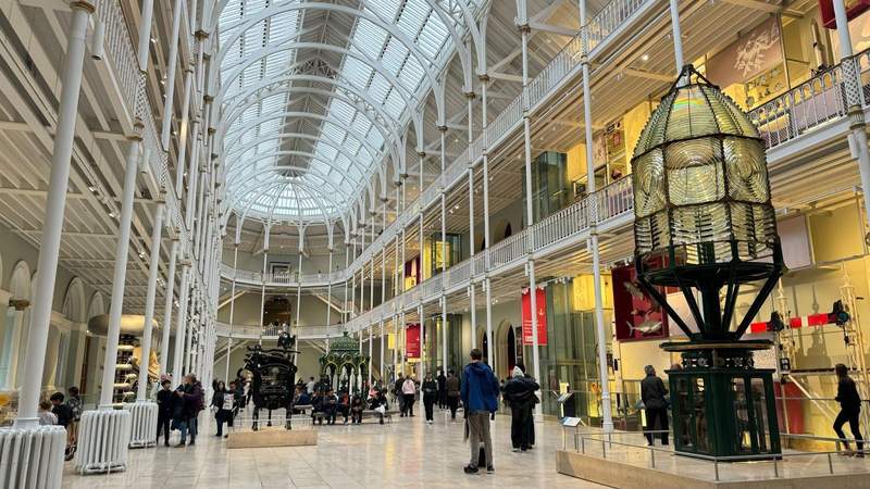 white interior with pillars large green glass machine and people walking around in the National Museum of Scotland