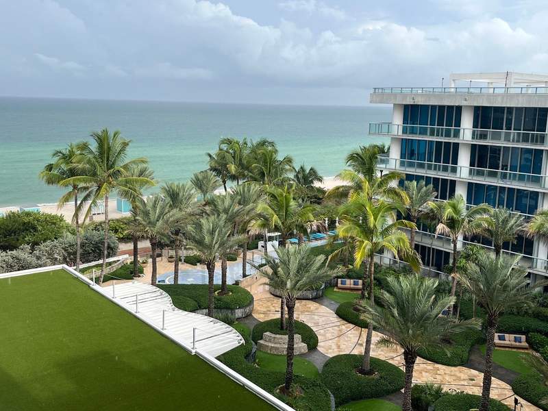 Palm trees a pool and a white resort with windows face the ocean. The sea waters are blue and green with gray clouds on the horizon.