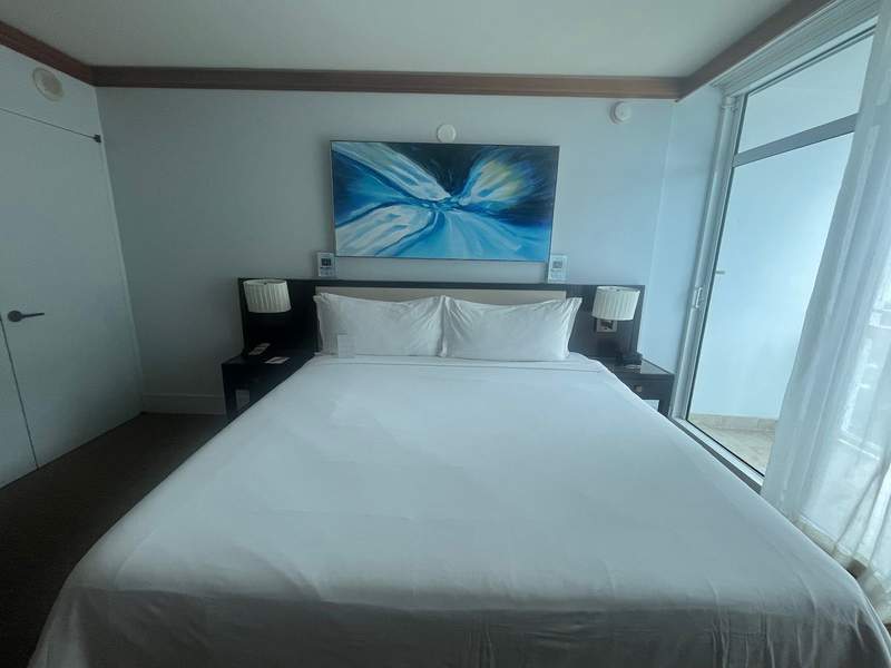 A king sized bed with white sheets. Light filters in through the patio and theres a blue painting hanging over the headboard.