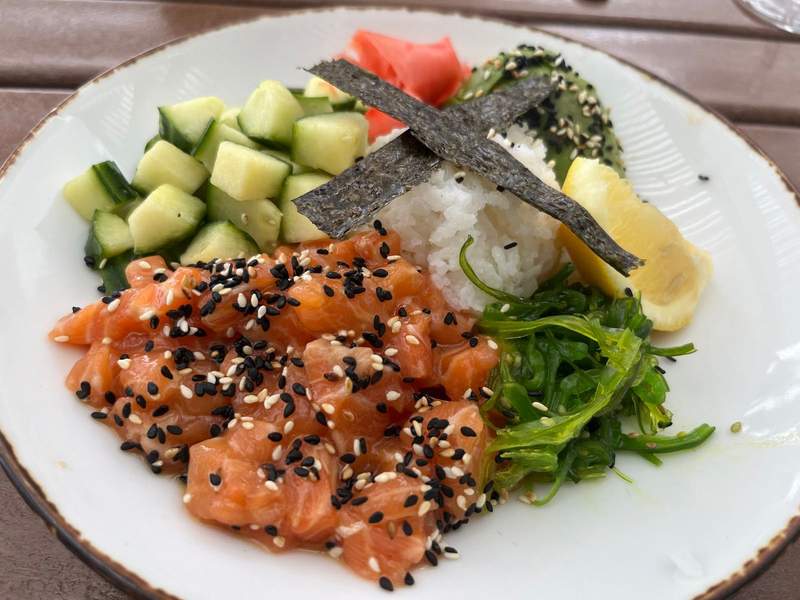 A salmon poke bowl containing pink raw fish cucumbers avocado white rice and seaweed salad is topped with crossed strips of nori.