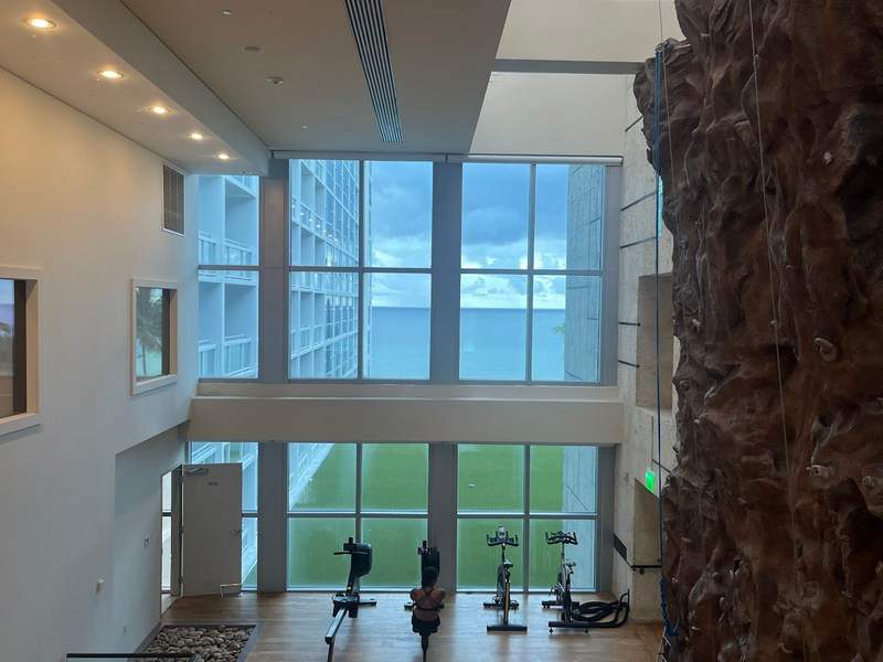 Four stationary bicycles look through soaring glass windows at the ocean below. A tall rock climbing wall is behind to the right.