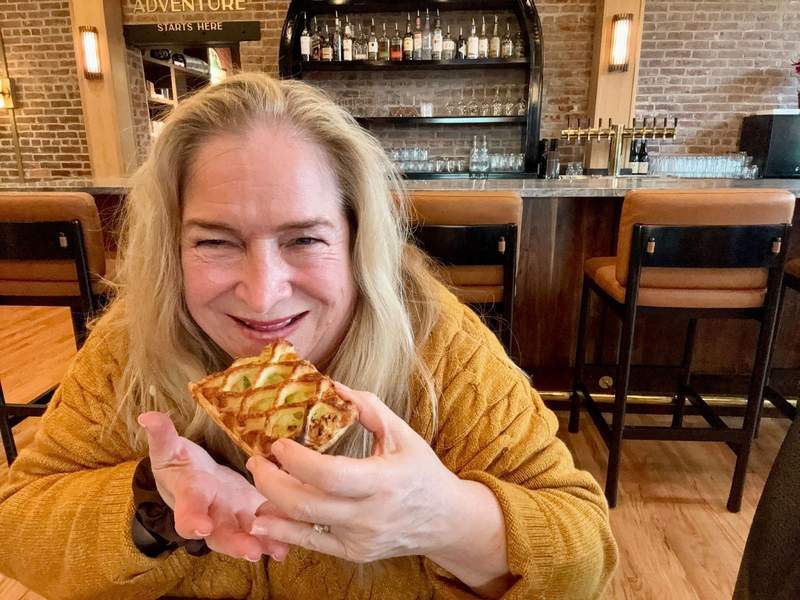 The author sits at a table with the lobby bar in the background eating a pastry. She is is wearing a gold sweater.