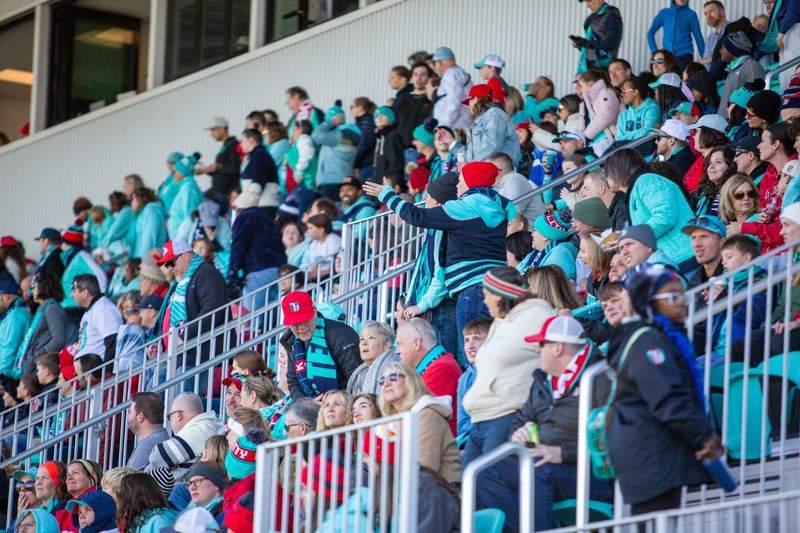 Rows of fans in teal blue support the FC Current soccer team.