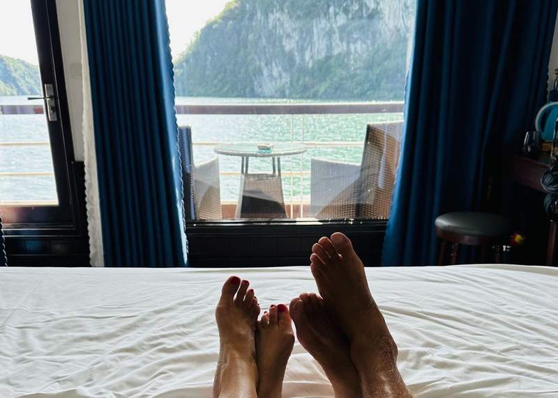 Feet resting on a bed while looking out open windows at limestone cliffs and water in Ha Long Bay