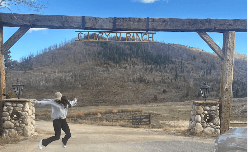 Woman jumping beneath the wooden entrance sign at C Lazy U Ranch in Colorado with rolling hills and open landscape in the background