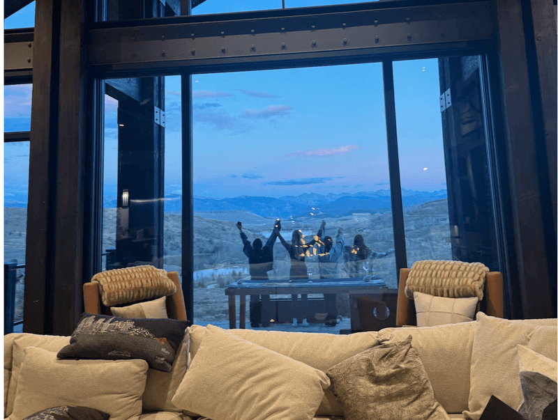 Group of people standing outside on a patio at dusk seen through large windows from inside a cozy lodge with mountain views beyond.
