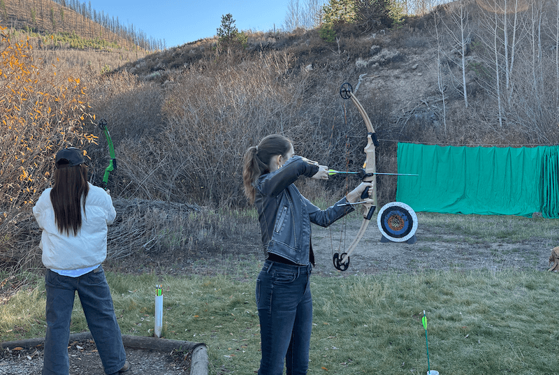 Woman aiming a bow and arrow at a target outdoors during an archery activity with another person practicing nearby.
