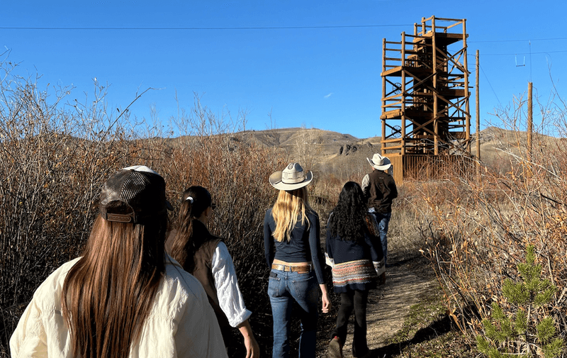 Group of women walking along a narrow trail toward a wooden observation tower in a dry brush filled landscape.