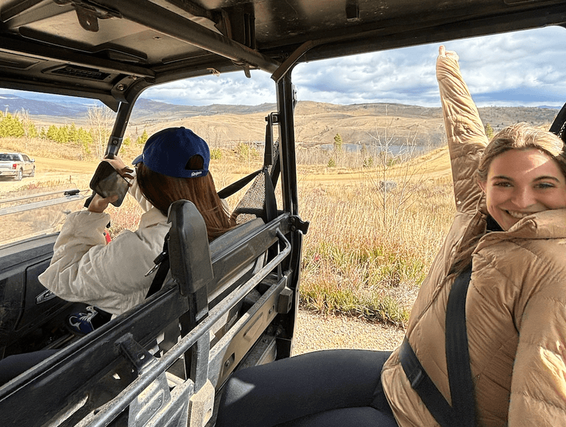 Two women riding in an open utility vehicle through grassy fields smiling and raising their arms as hills stretch beyond.