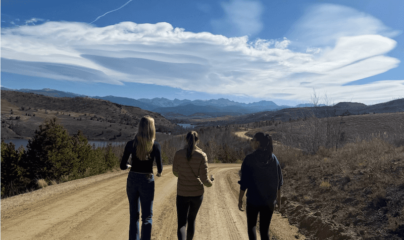 Three women walking along a dirt road through rolling hills and open countryside beneath a partly cloudy sky.