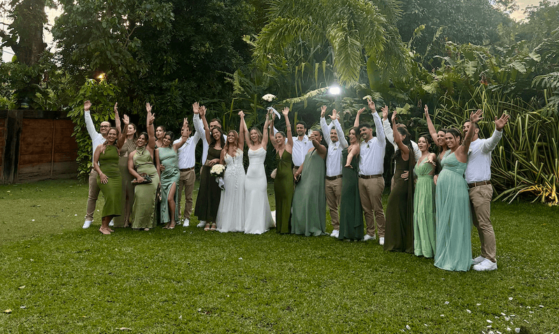 Group of wedding guests celebrating outdoors in Rio de Janeiro dressed in green and neutral tones with the bride and bride at the center raising their arms together.