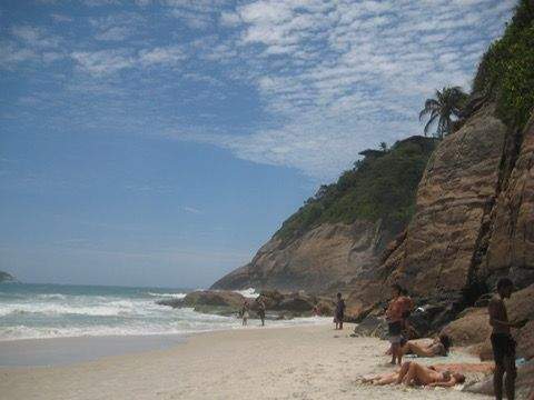 Small secluded beach in Rio de Janeiro with golden sand gentle waves and towering rocky cliffs where a few locals relax under a bright blue sky.
