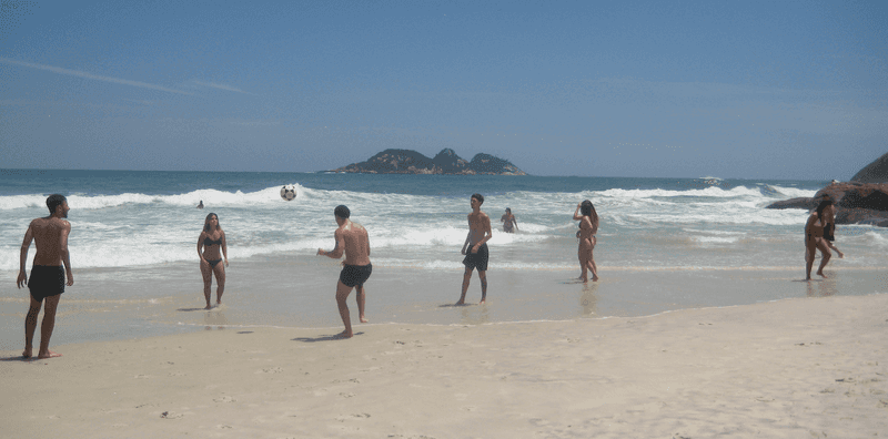 Group of people playing futevolei on a quiet Rio de Janeiro beach kicking a ball over a net using their feet with rocky cliffs and ocean waves in the background.