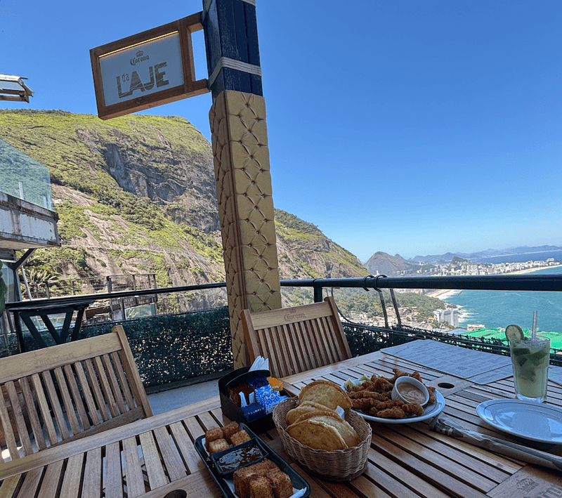 Rooftop dining table at La Laje in Rio de Janeiro with Brazilian appetizers and a caipirinha overlooking green mountains coastline and ocean on a sunny day.